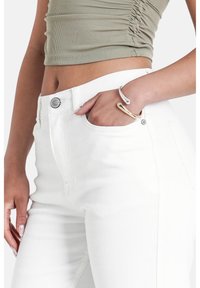 Modelo vistiendo una blusa verde oliva ajustada y jeans blancos, con la mano en el bolsillo, mostrando una pulsera de plata y oro contra un fondo neutro.