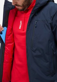 Navy waterproof jacket with red interior and blue phone visible in pocket. Features include zippered pockets and soft, textured lining.