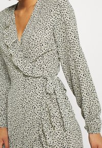 Robe grise avec un décolleté en V, des manches longues, des détails froncés et une taille ceinturée ; ornée d'un motif à pois noir et blanc.