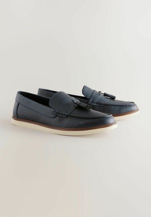 Mocassins en cuir bleu marine avec une languette texturée, semelle en caoutchouc blanc plat et détails de couture contrastés. Présente un accent en nœud à l'avant.