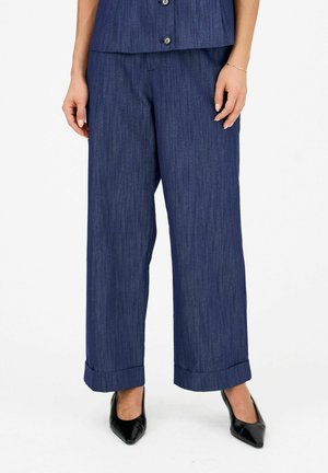 Pantaloni blu navy a gamba larga con orli risvoltati indossati con scarpe nere a punta, mostrati dalla vita ai piedi.