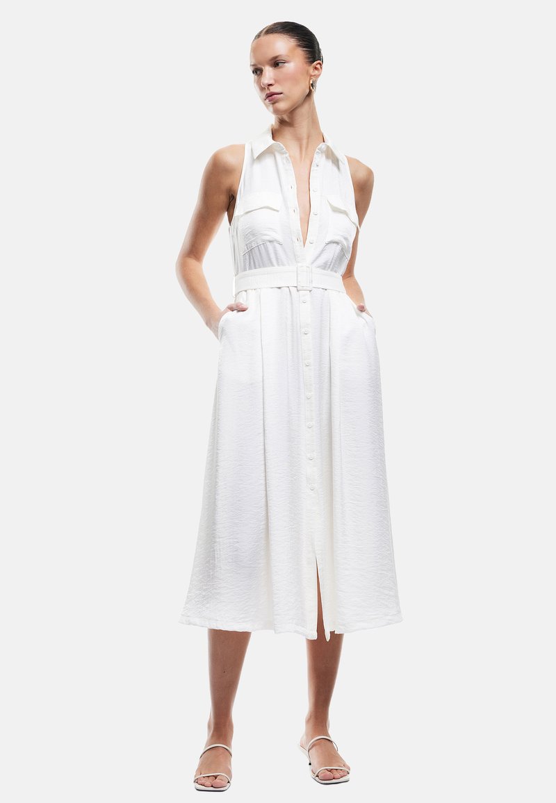 Vestido blanco sin mangas con botones en la parte delantera, cinturón en la cintura y bolsillos laterales. Presenta una tela texturizada y un corte a media longitud.
