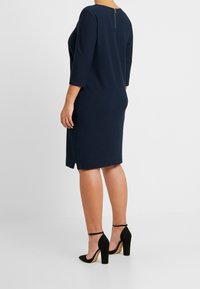 Robe bleu marine avec des manches 3/4, en tissu texturé, fermeture éclair au dos et fentes sur les côtés. Assortie à des chaussures à talons noirs.