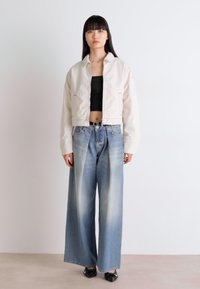 Lichtbeige cropped jack over een zwarte crop top, gecombineerd met wijduitlopende vervaagde blauwe spijkerbroek. Glanzende zwarte schoenen maken de outfit compleet.
