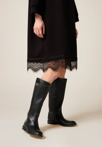 Bottes hautes en cuir noir avec une finition lisse, une couture blanche contrastante et une boucle décorative sur le côté.