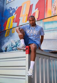 Lys blå grafisk t-skjorte med bandana mønster og "KANI" tekst, kombinert med denim shorts. Hvite sokker og grå joggesko. Vist mot et fargerikt veggmaleri.
