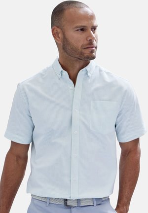 Mann mit kurzen Haaren und gepflegtem Bart, der ein hellblaues, kurzärmliges Hemd mit Knöpfen, Brusttasche und gemustertem Gürtel trägt.