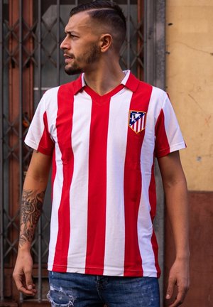 Rood-wit gestreept voetbalshirt met een V-hals, met een schildlogo op de linkerborst; gedragen met versleten blauwe jeans.