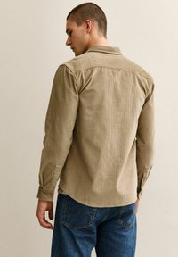 Mężczyzna odwrócony plecami, ubrany w Beżową, teksturowaną koszulę z długim rękawem oraz niebieskie dżinsy, stojący przy gładkiej, jasnej ścianie.