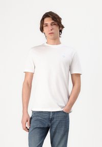 T-shirt bianco in cotone con maniche corte, colletto rotondo e un logo azzurro sul lato sinistro del petto. Indossato con jeans in denim blu.