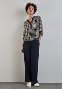 Camicetta a maniche lunghe con motivo geometrico bianco e nero, con scollatura a V, abbinata a pantaloni navy a gamba larga e sneaker bianche, su uno sfondo neutro.