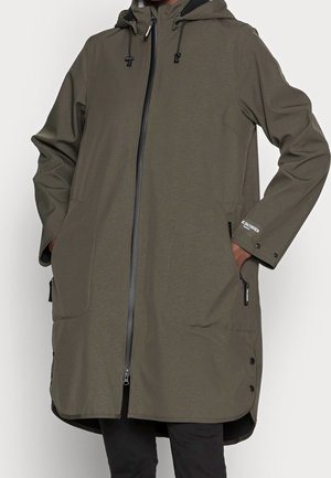 Personne portant un long manteau à capuche vert olive avec fermeture éclair à l'avant et les mains dans les poches latérales, debout devant un fond uni.