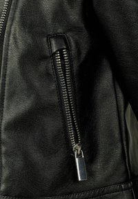 Dettaglio di una giacca in finta pelle nera con una tasca con cerniera dotata di hardware argentato e superficie testurizzata. Focus sulla cucitura e sul design della cerniera.