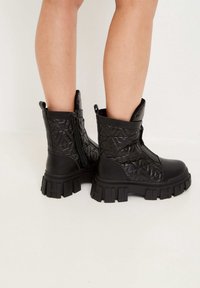 Botines negros hasta el tobillo con parte superior de cuero acolchado, suelas gruesas de goma, cremalleras laterales y tiradores. Diseño texturizado con un patrón geométrico.