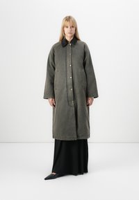 Cappotto di jeans verde oversize con colletto nero, dotato di grandi tasche e bottoni frontali, indossato sopra un abito nero lungo fino ai piedi.