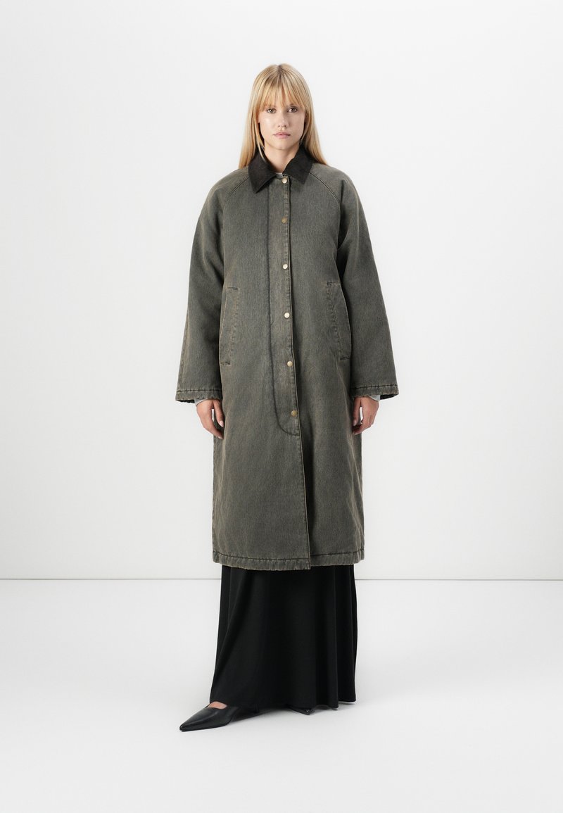 Cappotto di jeans verde oversize con colletto nero, dotato di grandi tasche e bottoni frontali, indossato sopra un abito nero lungo fino ai piedi.