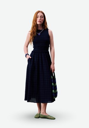 Femme aux longs cheveux roux portant une robe midi à carreaux bleu marine sans manches, des ballerines en filet vert, un collier, un bracelet, et tenant un sac rayé bleu-vert.