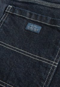 Donkerdenimstof met contrasterende witte stiksels. Bevat een label met de tekst "STANDARD ISSUE" op een pocket met zichtbare textuur en nerf.