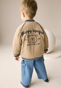 Jasnoszara kurtka z kordurowego materiału z granatowymi akcentami; na plecach nadruk z grafiką niedźwiedzia i napisem "Happy League". Połączona z niebieskimi spodniami cargo z denimu.