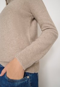 Beżowy sweter zdzierany z długim rękawem o prążkowanym wykończeniu, charakteryzujący się miękką fakturą. W komplecie z niebieskimi dżinsami z detalem w postaci kieszeni.