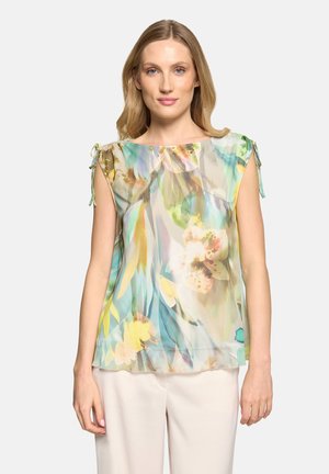 Vrouw draagt een mouwloze semi-transparante bloemenblouse met schouderstrikjes en lichtgekleurde broek, staand tegen een effen witte achtergrond.