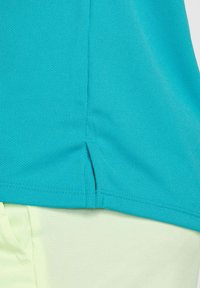 Top deportivo turquesa con un tejido texturizado, con una abertura lateral en el dobladillo, combinado con pantalones de un verde claro.