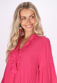 Bluză roz din material texturat, cu decolteu în V, accente cu ciucuri și mâneci evazate. Modelul poartă cercei colorati și lungi.