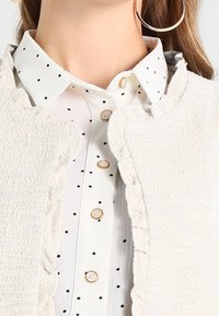 Biały teksturowany kamizelka z falistymi krawędziami na koszuli z kołnierzykiem, zdobionej czarnymi kropkami i złotymi guzikami. Widoczne duże kolczyki koła.