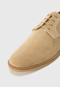 Zapato oxford de ante beige con punta redondeada, textura suave y cierre de cordones con cinco ojetes, acentuado por una suela marrón clara.