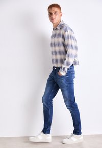 Camisa de cuadros en azul y beige, de mangas largas, combinada con jeans azul oscuro y zapatillas blancas. Ajuste casual con cuello abotonado.