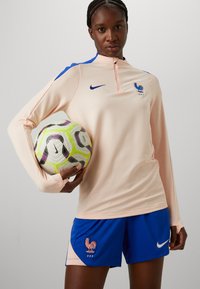 Nike sportinė apranga, kurią sudaro šviesiai rožinė ilgomis rankovėmis palaidinė su mėlynomis detalėmis ir Prancūzijos futbolo federacijos logotipu, derinama su mėlynomis šortais. Laiko raštuotą futbolo kamuolį.