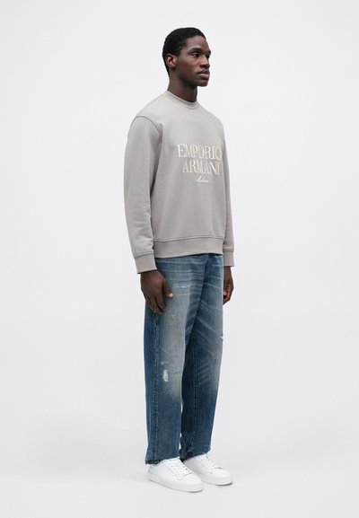 Šedá mikina so logom "Emporio Armani", skombinovaná so svetlomodrými roztrhnutými džínsy a bielymi teniskami. Model stojí proti jednofarebnému pozadiu.