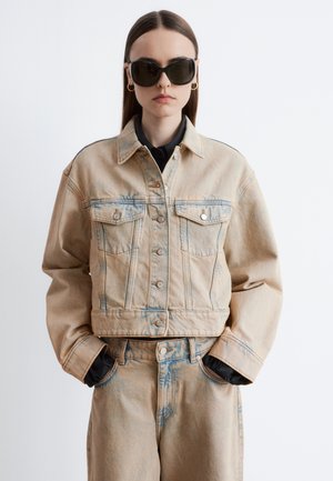 Kvinde iført beige denimjakke og bukser, sort skjorte, store sorte solbriller og guldringe med hænderne i lommerne.