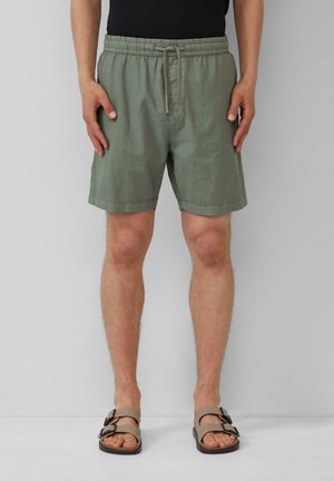 Man draagt olijfgroene shorts met trekkoord en beige sandalen met gesp, staand tegen een grijze muur.