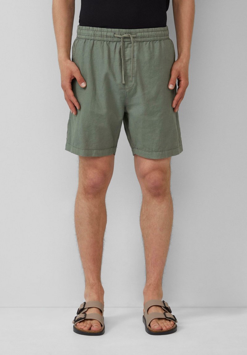 Homme portant un short vert olive à cordon et des sandales beiges à boucle, debout contre un mur gris.