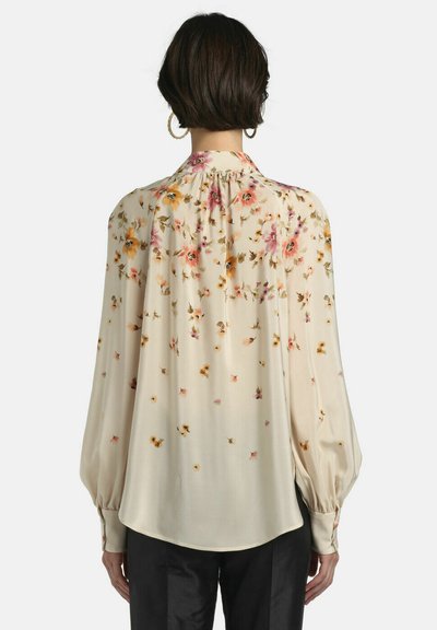 Woman with short dark hair wearing large hoop earrings and a cream floral blouse with long puffed sleeves viewed from the back.