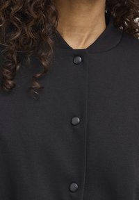 Czarna kurtka na guziki z prążkowanym kołnierzem, wykonana z gładkiej tkaniny, z zaokrąglonymi czarnymi guzikami na przodzie.