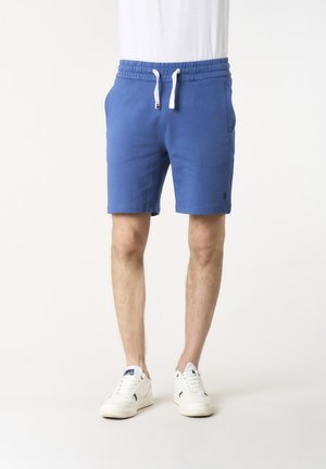 Uomo che indossa pantaloncini blu con coulisse e scarpe da ginnastica bianche, in piedi su uno sfondo bianco semplice.