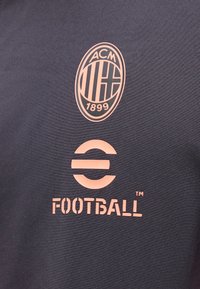 Λογότυπο AC Milan πάνω από ένα στυλιζαρισμένο λογότυπο "E FOOTBALL" σε πορτοκαλί σε σκοτεινό ύφασμα, πιθανώς αθλητική φανέλα ή ένδυμα.