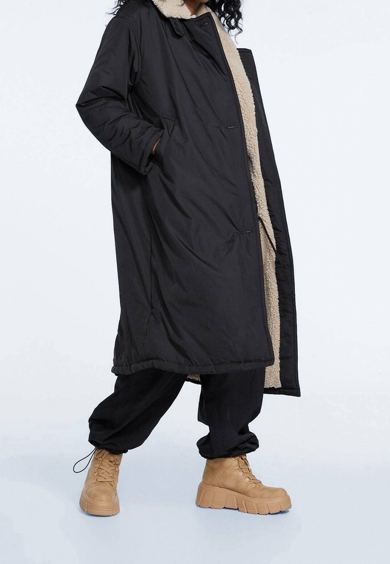 Personne portant un long manteau noir avec une doublure en polaire beige, un pantalon noir et des bottes beige à lacets épaisses, debout avec une main dans la poche du manteau.