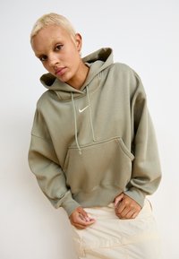 Hoodie oversized em verde-oliva com um bolso frontal e capuz com cordão, apresentando um pequeno logo branco da Nike no peito. Textura de tecido macio.