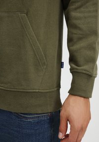Olive green sweatshirt featuring a kangaroo pocket, ribbed cuffs, and hem, with a small label on the side. Worn over denim jeans.