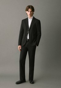 Traje negro a medida con un corte ajustado, blazer de un solo botón, camisa de vestir blanca y zapatos de vestir negros. Tejido suave, detalles mínimos.