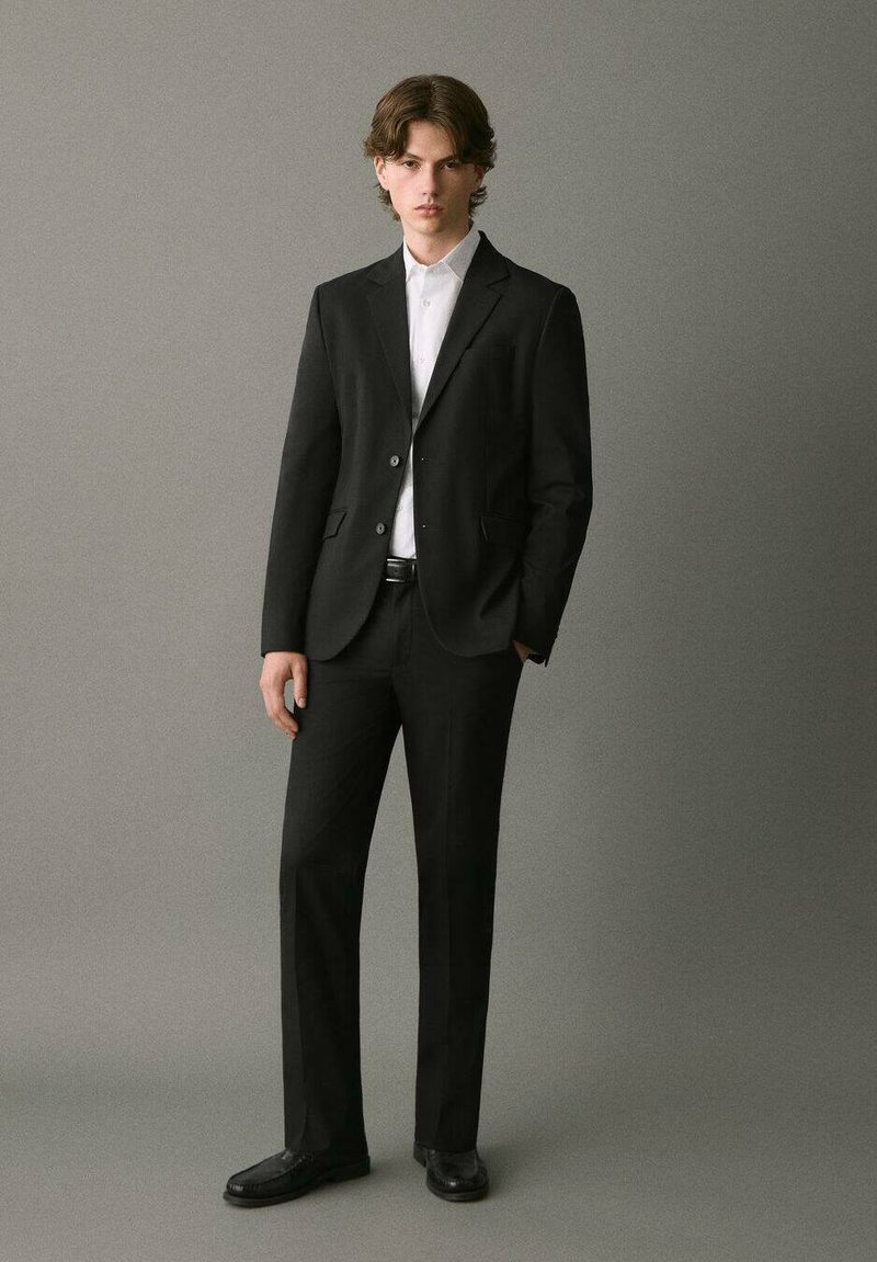 Traje negro a medida con un corte ajustado, blazer de un solo botón, camisa de vestir blanca y zapatos de vestir negros. Tejido suave, detalles mínimos.