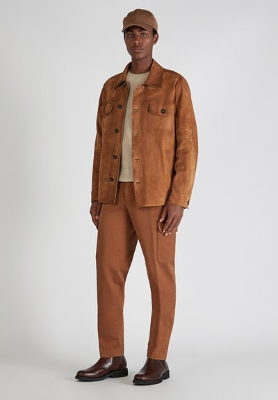 Bruine suède jas met twee borstzakken, beige gebreide trui, bruine cargobroek en bruine leren Chelsea boots. Pet past bij de kleur van de jas.