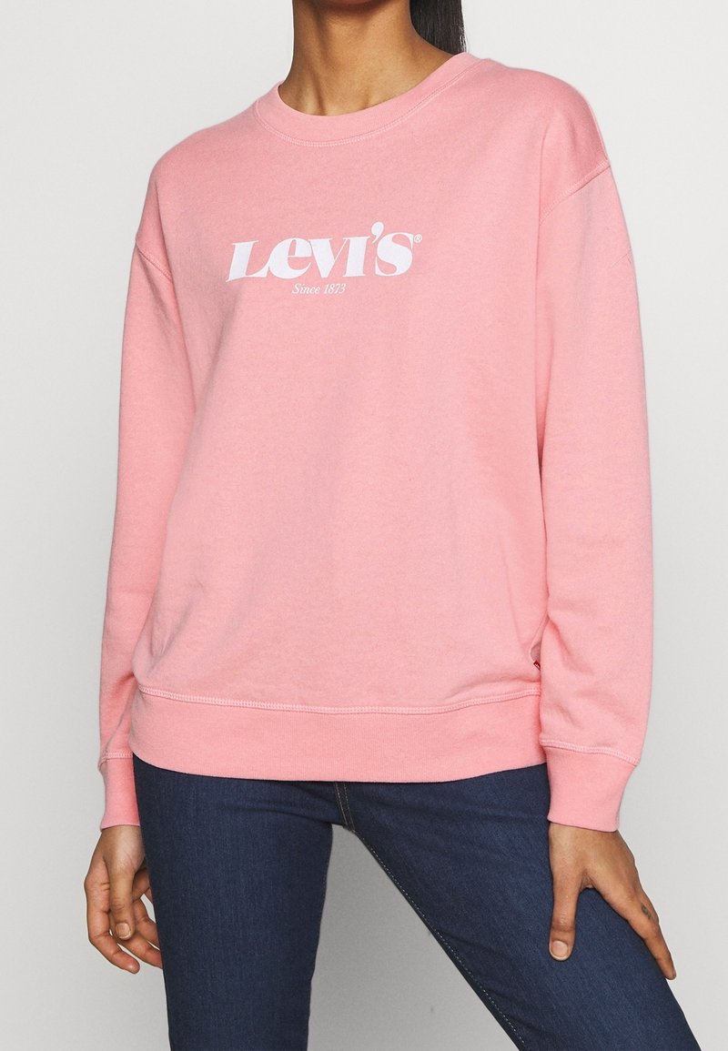 Femme portant un sweat-shirt Levi's rose avec un logo texte blanc, associée à un jean bleu foncé, debout devant un fond gris clair uni.