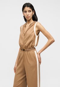 Ολόσωμη φόρμα με κουκούλα, χωρίς μανίκια, σε ανοιχτό καφέ χρώμα με αντίθετες κρεμ ρίγες στα πλάγια. Διαθέτει χρυστό φερμουάρ, ζώνη και άνετη εφαρμογή. Μαλακή υφή υφάσματος.