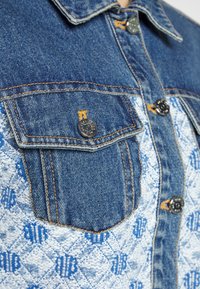 Blå denimjacka med ett kontrasterande mönstrat tyg. Den har två bröstfickor, svarta knappar och detaljer i orange sömnad.