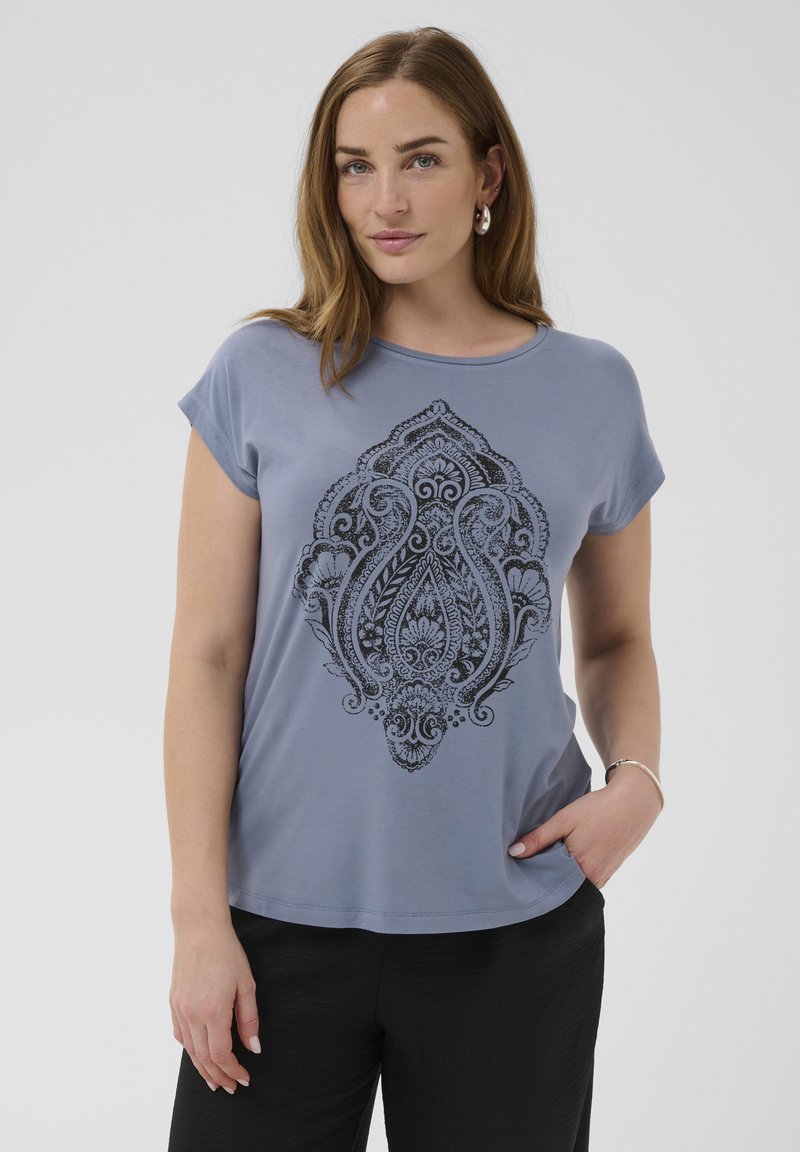 Femme aux cheveux châtain clair portant une chemise bleue à manches courtes avec un motif floral et cachemire noir complexe, un pantalon noir et des bijoux en argent.