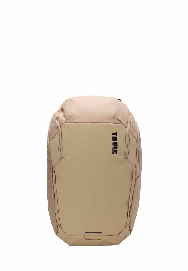 Sac à dos beige de forme arrondie, fabriqué en tissu résistant, arborant un logo noir, une fermeture éclair et un panneau avant texturé pour une meilleure prise en main.